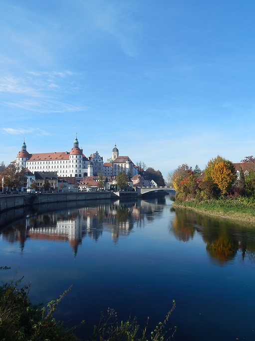 Neuburg an der Donau. Im Vordergrund die Donau. In der Mitte das Schloss, das sich im Fluss spiegelt. Blauer Himmel.