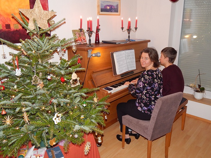 Links ein Weihnachtsbaum, rechts spielt eine Mama mit ihrem Sohn 4-händig Klavier. 